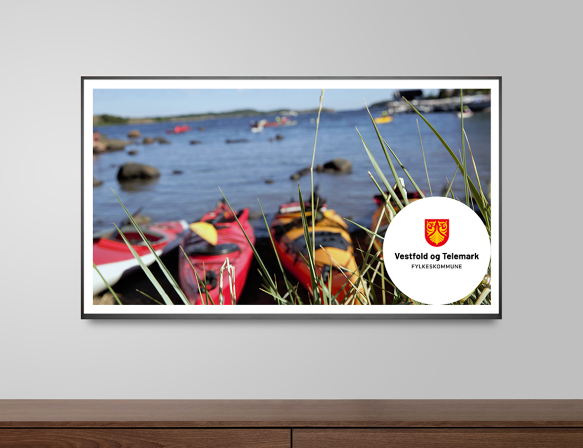 Vegghengt tv-skjerm med bilde av hovedlogo plassert på et foto av kajakker liggende i strandkanten